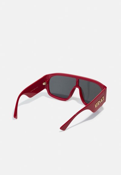 Ochelari de soare din plastic roșu cu lentile dreptunghiulare, nuanță gri închis și logo-ul "VERSACE" în aur pe laterale. Suprafață netedă, design îndrăzneț.