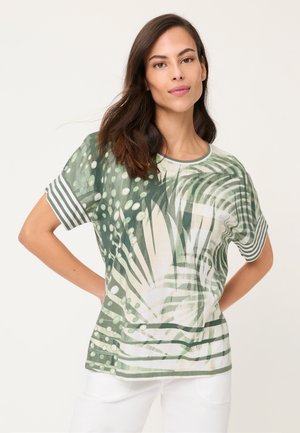 Frau mit langen dunklen Haaren, die ein grün-weiß gemustertes kurzärmeliges Hemd und weiße Hose trägt, steht mit den Händen in der Hüfte.