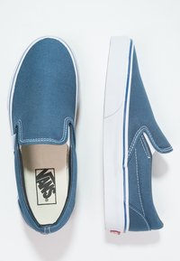 Blå canvas slip-on skor med vita gummisulor, kontrasterande sömmar och en logo på innersulan. Enkelt design utan snören.