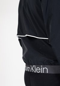 Giacca a vento nera con bordi bianchi, caratterizzata da un taglio corto e una fascia elastica con logo Calvin Klein. Texture liscia e design minimal.