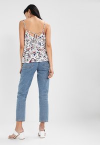 Donna che indossa un top senza maniche bianco con fiori e nodo sulla schiena, jeans blu, sandali bianchi con tacco a blocco e orecchini a cerchio dorati, in piedi di spalle.
