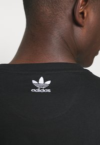 Svart bomullströja med rund hals, prydd med en vit broderad Adidas trefoil-logotyp på baksidan nära kragen.