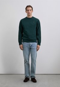 Homme adossé à un mur uni, portant un sweat à capuche vert foncé, un jean bleu clair et des chaussures à lacets marron foncé.