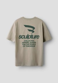 T-shirt en coton beige à manches courtes et col rond. Présente un texte vert foncé : "sculpture" et "COLLECTIVE APPAREL EDITION FOR THE PEOPLE OF TOMORROW."