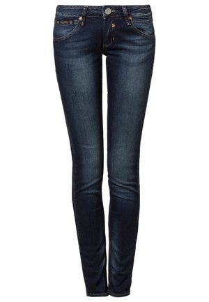 Vrouw loopt met een roze en zwarte gestreepte top met lange mouwen, donkere skinny jeans en zwarte schoenen met studs tegen een effen achtergrond.