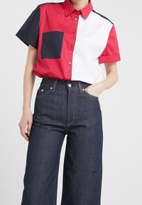 Camisa de manga corta en bloques de color rojo, blanco y marino con un bolsillo frontal. Pantalones vaqueros oscuros de talle alto y pernera ancha.
