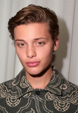 Persoană tânără cu păr ondulat și castaniu, ochi caprui deschis, purtând o jachetă verde cu model, cu guler și nasture, expresie neutră.