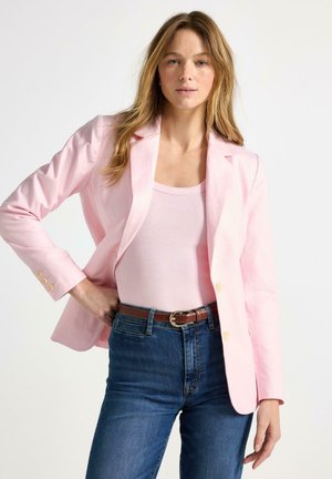 Kvinde iført lyserød blazer, matchende tanktop og blå jeans med brunt bælte, poserer med den ene hånd på hoften mod en ensfarvet baggrund.
