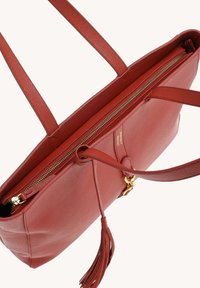 Sac en cuir rouge avec fermeture éclair dorée, longues poignées et un gland décoratif sur le fermoir de la poche avant.