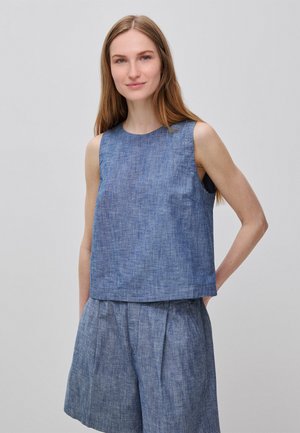 Jeune femme aux longs cheveux châtain clair portant un haut bleu sans manches et un short taille haute assorti sur un fond clair uni.