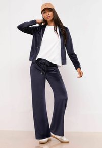 Jūras spēku velūra treniņtērps ar rāvējslēdzēja kapuci un platām biksēm. Baltā T-kreklā zemā un bēšā cepurē ar "NY" uz priekšpuses. Baltas sporta kurpes.