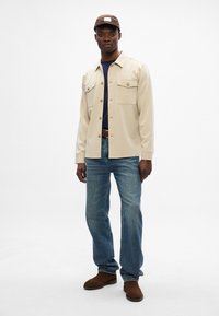 Μπέζ πουκάμισο με κουμπιά και δύο τσέπες στο στήθος, συνδυασμένο με ανοιχτό μπλε τζιν και καφέ σουέτ μποτάκια. Μοντέλο που φορά καφέ καπέλο.