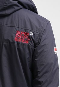 Veste bleu marine foncé avec capuche, avec le texte brodé en rouge "SUPER DRY JPN" et des caractères japonais sur la partie arrière de l'épaule.