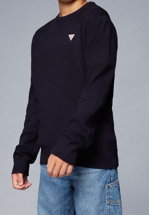 Niño vistiendo un suéter negro de manga larga con un pequeño logo triangular en el pecho y jeans de mezclilla azul claro con un bolsillo lateral.