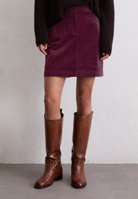 Jupe courte en velours côtelé bordeaux avec poche avant et fermeture à bouton, associée à des bottes en cuir marron arrivant au genou, au fini lisse et avec une boucle discrète.