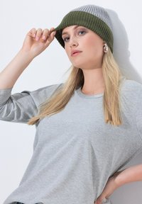 Gorro de punto en gris con una banda verde oscura, llevado inclinado hacia atrás; combinado con una camiseta de manga larga gris claro con cuello acanalado.