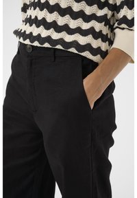 Pantaloni in lino neri con una texture liscia, dotati di tasche laterali e chiusura a bottone, abbinati a un maglione in maglia a fantasia.