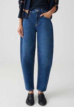 Persona che indossa jeans blu larghi, mocassini neri e una maglia blu navy con una mano in tasca, in piedi su uno sfondo chiaro e semplice.