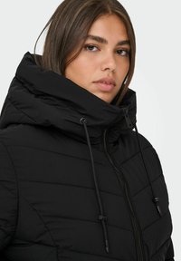 Chaqueta acolchada negra con un alto cuello, que presenta secciones acolchadas, cordones ajustables y un zipper frontal. Acabado liso y mate.