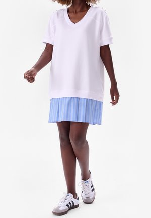 Woman wearing white v-neck oversized T-shirt, blue and white vertical striped skirt, and white Adidas sneakers with black stripes walking.