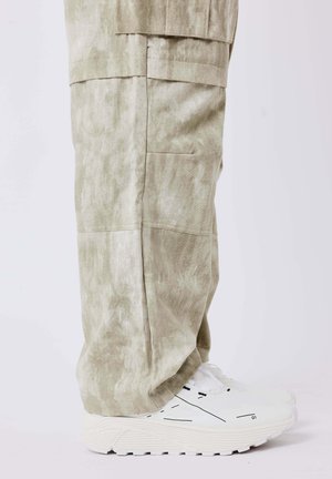 Pantalones verde claro con textura y bolsillos laterales, combinados con zapatillas blancas que tienen detalles en negro y suelas gruesas.