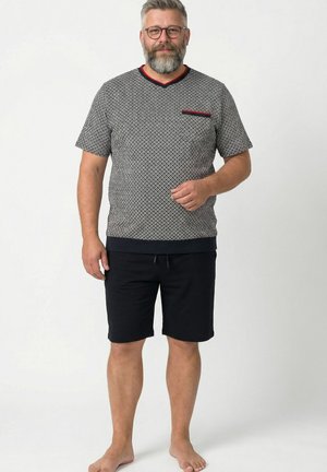 Mand med gråt skæg og briller iført mønstret kortærmet skjorte med rød kant og sorte shorts står barfodet på ensfarvet baggrund.