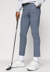 Zīmēti bikses ar zilu ģeometrisku dizainu. Izgatavotas no viegla auduma, tās ir kombinētas ar baltām sporta kurpēm un ikdienišķu pelēku džemperi.