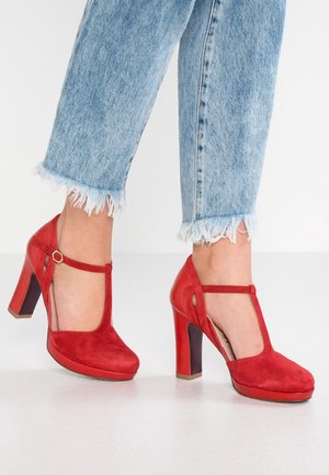 Voeten in lichtblauwe, versleten jeans met een rafelrand, gekleed in rode suède T-strap hakken met een blokhak, staande op een witte vloer.