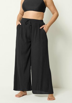 Kvinde iført løse sorte bukser med vidde og elastisk talje med snøre, kombineret med en sort crop top, stående barfodet.