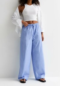 Vrouw in een witte mouwloze crop top, losse lichtblauwe drawstringbroek, witte open shirt en zwarte slippers, staand tegen een witte muur.