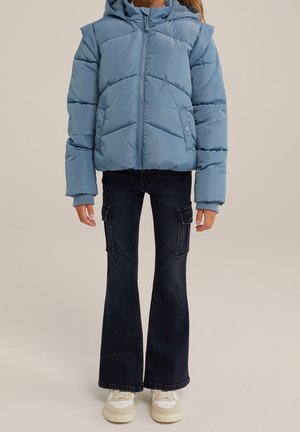 Kind trägt eine hellblaue gesteppte Kapuzenjacke, schwarze ausgestellte Cargo-Jeans und weiße Turnschuhe und steht vor einem beigen Hintergrund.