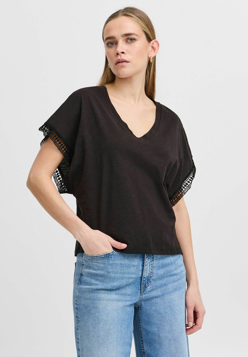 Sort v-hals top med korte dolmanærmer, der har blondekant. Blødt materiale, afslappet pasform, parret med lyseblå denimjeans.
