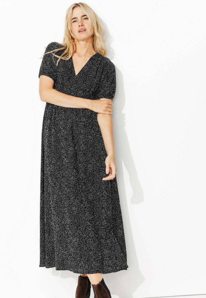 Robe maxi noire avec des éclats blancs, décolleté en V, manches courtes bouffantes et une silhouette fluide. Associée à des bottes sombres, longueur cheville.