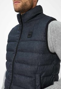 Dunkelblaues, gesteppte Weste mit hohem Kragen, Frontreißverschluss und schwarzem Stoffetikett. Unter den Ärmeln sind Strickärmel sichtbar. Texturiertes Material.