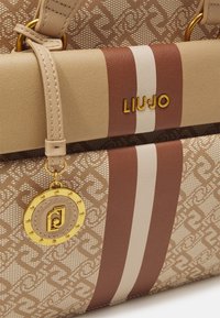 Beżowa torebka z wzorzystego materiału. Posiada brązowe i białe paski, złote akcenty oraz zawieszkę o okrągłym kształcie.