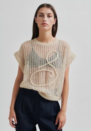 Strikket kortermet genser i beige med ribbestruktur og store løkkedetaljer. Kombinert med mørk denim høytlivsbukser.