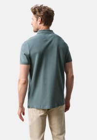 Pikétröja i dämpad teal-färg, korta ärmar och klassisk krage. Har sidokilar vid nederkanten för komfort och rörelsefrihet.