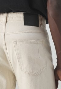 Vaqueros de mezclilla beige con un corte de tiro medio. Presentan un parche de marca de cuero negro y bolsillos traseros clásicos con costuras detalladas.