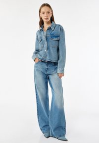 Chaqueta de mezclilla de color azul claro con dos bolsillos en el pecho y cierre de botones, combinada con unos pantalones anchos de mezclilla azul claro que presentan una textura deslavada.