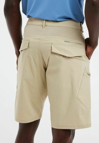 Beige cargoshorts hebben een gladde textuur, een elastische tailleband en twee achterzakken met kleppen. De merknaam "PROTEST HIGH PERFORMANCE" is zichtbaar.