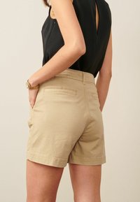 Beige Baumwollshorts mit Seitentaschen, flacher Vorderseite und kontrastierenden Nähten. Getragen mit einem schwarzen Oberteil; Seitenansicht mit Fokus auf die Details des Bunds.