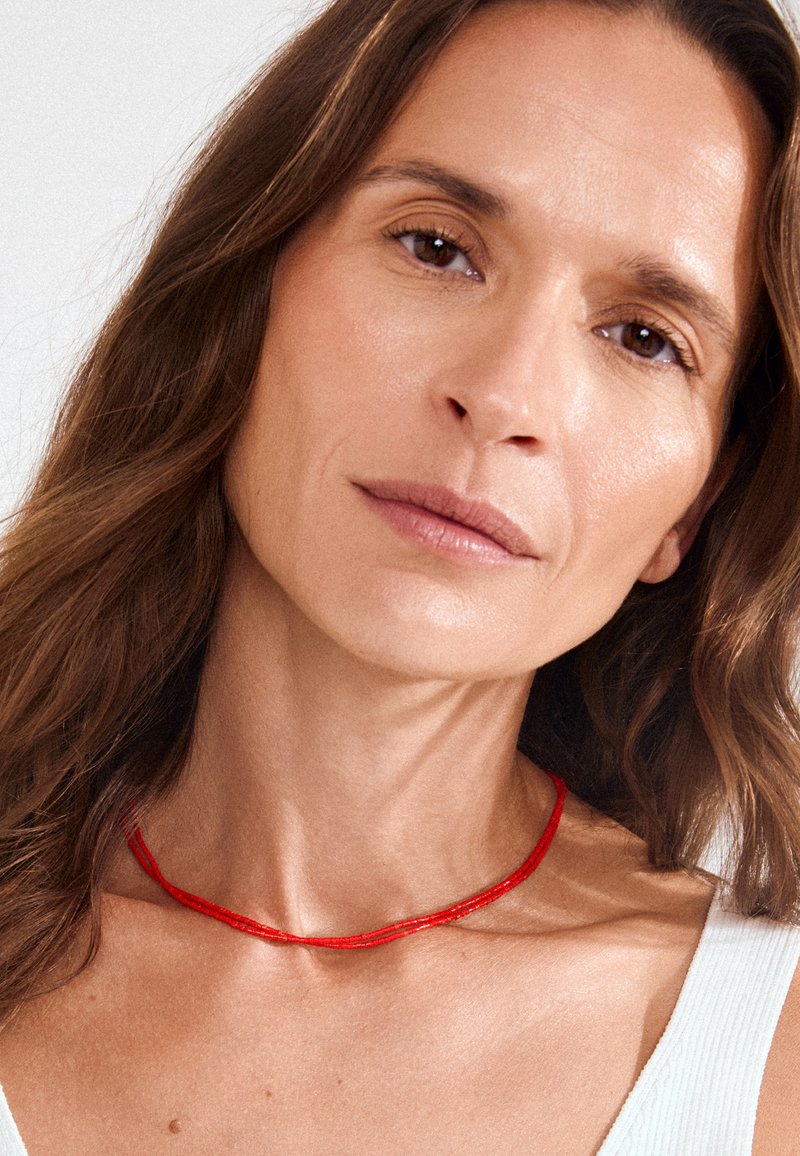Mujer con cabello largo y castaño, lleva un collar rojo fino y una camiseta sin mangas blanca, mirando directamente a la cámara con una expresión neutral.