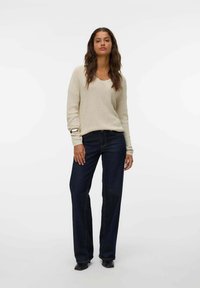 Maglione a maglia beige chiaro con scollo a V, abbinato a jeans a gamba larga blu scuro. L'outfit presenta delicati dettagli sulle maniche e una vestibilità rilassata.