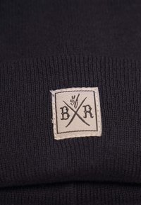 Svart stickad mössa med en krämfärgad fyrkantig etikett som har "B/R" och korslagda linjer, vilket visar på en mjuk textur och en åtsittande design.