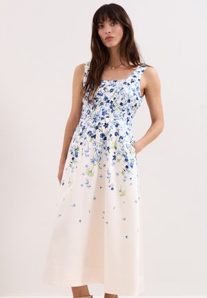 Femme aux longs cheveux foncés portant une robe blanche sans manches à motif floral bleu, debout devant un fond uni avec une main dans la poche.