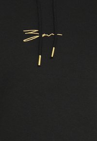 Sudadera negra con un logo bordado en dorado. Cuerdas gruesas con puntas metálicas doradas. Textura suave, diseño casual y estilo minimalista.