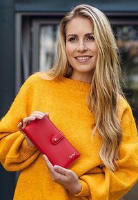 Red leather wallet with a snap closure, featuring a smooth texture and embossed logo, held in hands. Warm yellow sweater background.