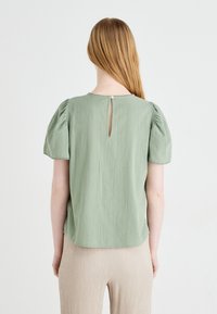 Svetlo zelena bluzica iz teksturirane tkanine. Ima balonske rukave, otvor v obliki ključavnice na hrbtu in zaobljen rob.