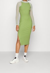 Nainen, joka on pukeutunut vihreään ribattu midimekkoon, jossa on kyljessä halkio, läpinäkyvään pitkähihaiseen ruudulliseen puseroon, valkoisiin sukkiin ja valkoisiin tennareihin, seisoo harmaalla lattialla.