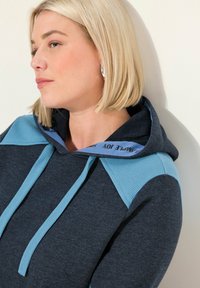 Felpa con cappuccio di un blu scuro, caratterizzata da un accento in mesh azzurro chiaro sulle spalle, cordini abbinati e un cappuccio con un colore interno in contrasto.